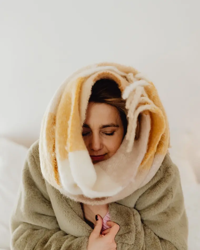 A woman wrapped in a blanket and a heavy coat indoors, eyes closed, trying to stay warm.