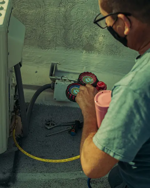 A technician in glasses reading a refrigerant gauge set connected to an outdoor unit.